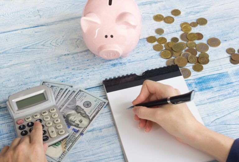 a white woman writes on white paper with a calculator next to her and a pink piggy bank in front of her.  Setting a budget is crucial in planning a road trip.