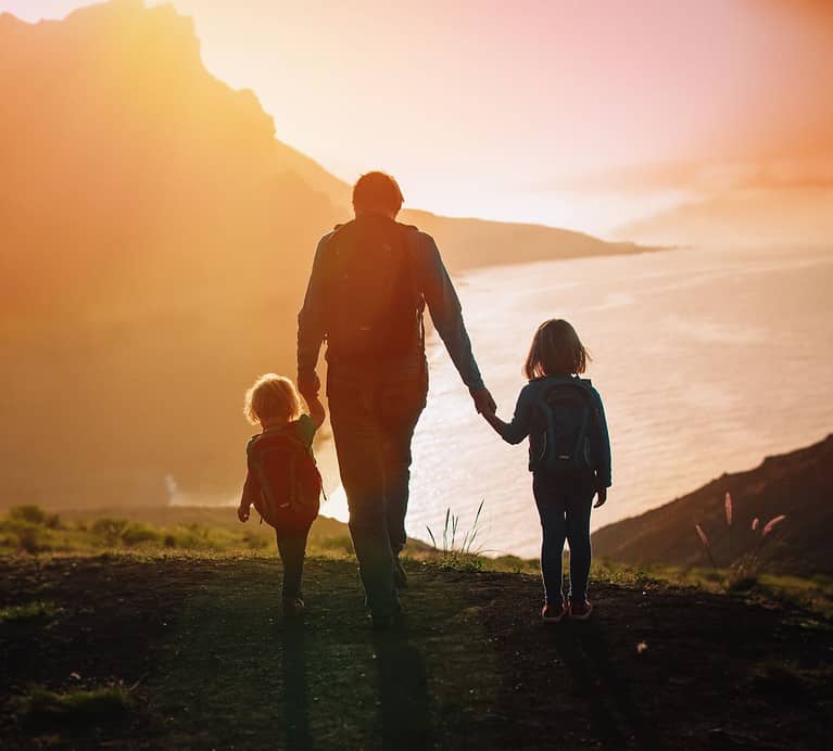 A parent holds the hands of his two young children as they walk toward the ocean.  Traveling with family can reduce stress and is just one reason why travel is important