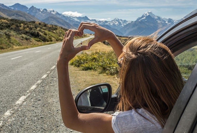 A blonde-haired white woman leans out of her car alongside a road and makes a heart shape with her hands in front of a mountain range.