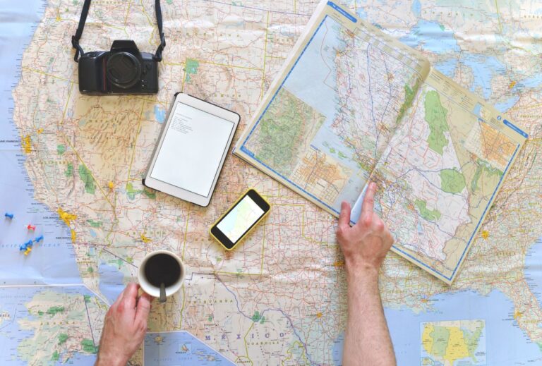 A road atlas is open and sits on a map of the United States.  Also on the map are a cell phone, a tablet, and a black camera.  A road atlas is an essential tool for any accessible road trip.