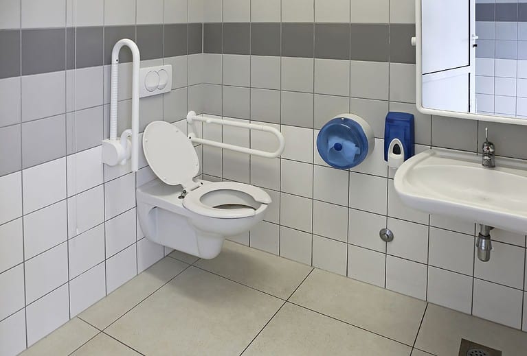 A white and grey wheelchair accessible bathroom has a toilet with two fold down grab bars next to it and a roll under sink.  Knowing where accessible restrooms are is crucial for any road trip.