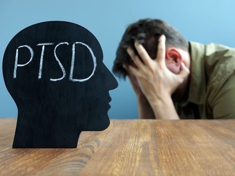 a white man with dark hair sits with his head in his hands.  On the table in front of him is a black head with the letters PTSD on it.