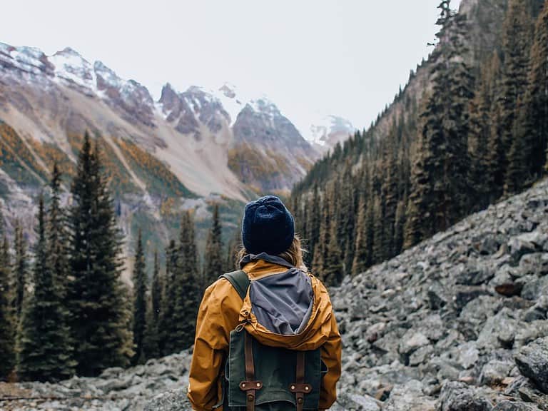 A person with long blonde hair wears a blue hat and yellow jacket and is admiring the forest and mountains in the background.  Hiking and being out in nature is a great trip for people traveling with PTSD.