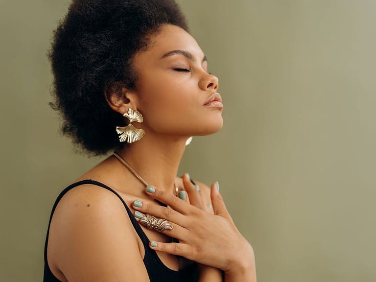 a black woman is standing with her eyes closed and her hands over her heart as she practices being mindful of her body.  