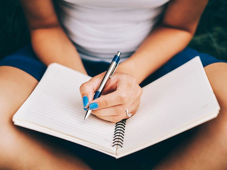 A white woman is sitting with crossed legs and writing in a journal.  Journaling is a great coping skill for people traveling with PTSD.