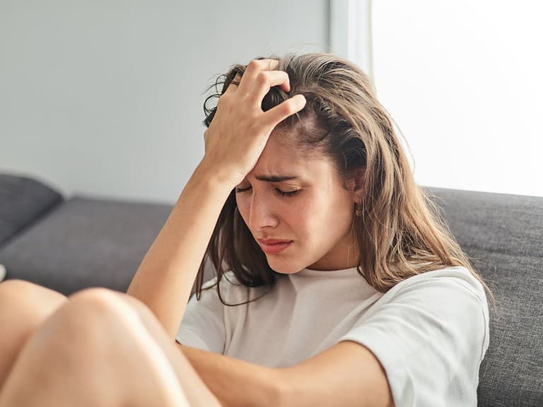 A white woman with brunette hair is sitting on a grey couch with her hand on her head and she looks distressed.