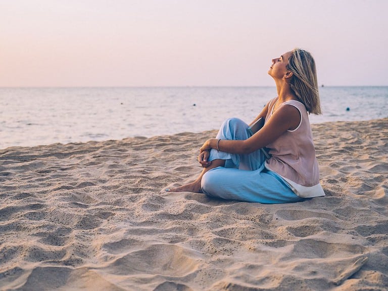 An older woman sits on a sandy beach and breathes in the ocean air as she relaxes.