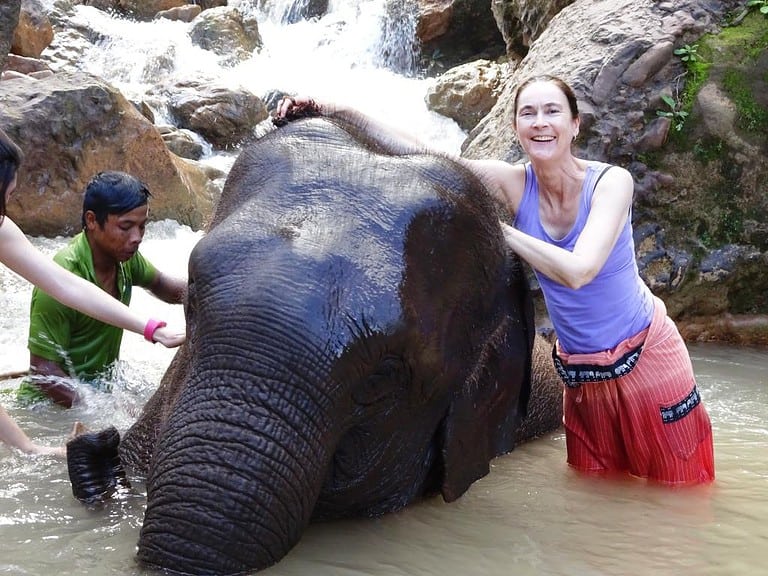 A white woman with dark hair stands in murky waters besides an Asian elephant.  Moments like this are how traveling the world can change your personality by helping you become more mindful.