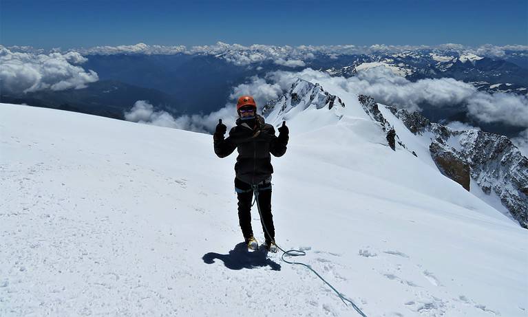 A woman in a black snow suit and orange helmet stands on top of a mountain in France.  