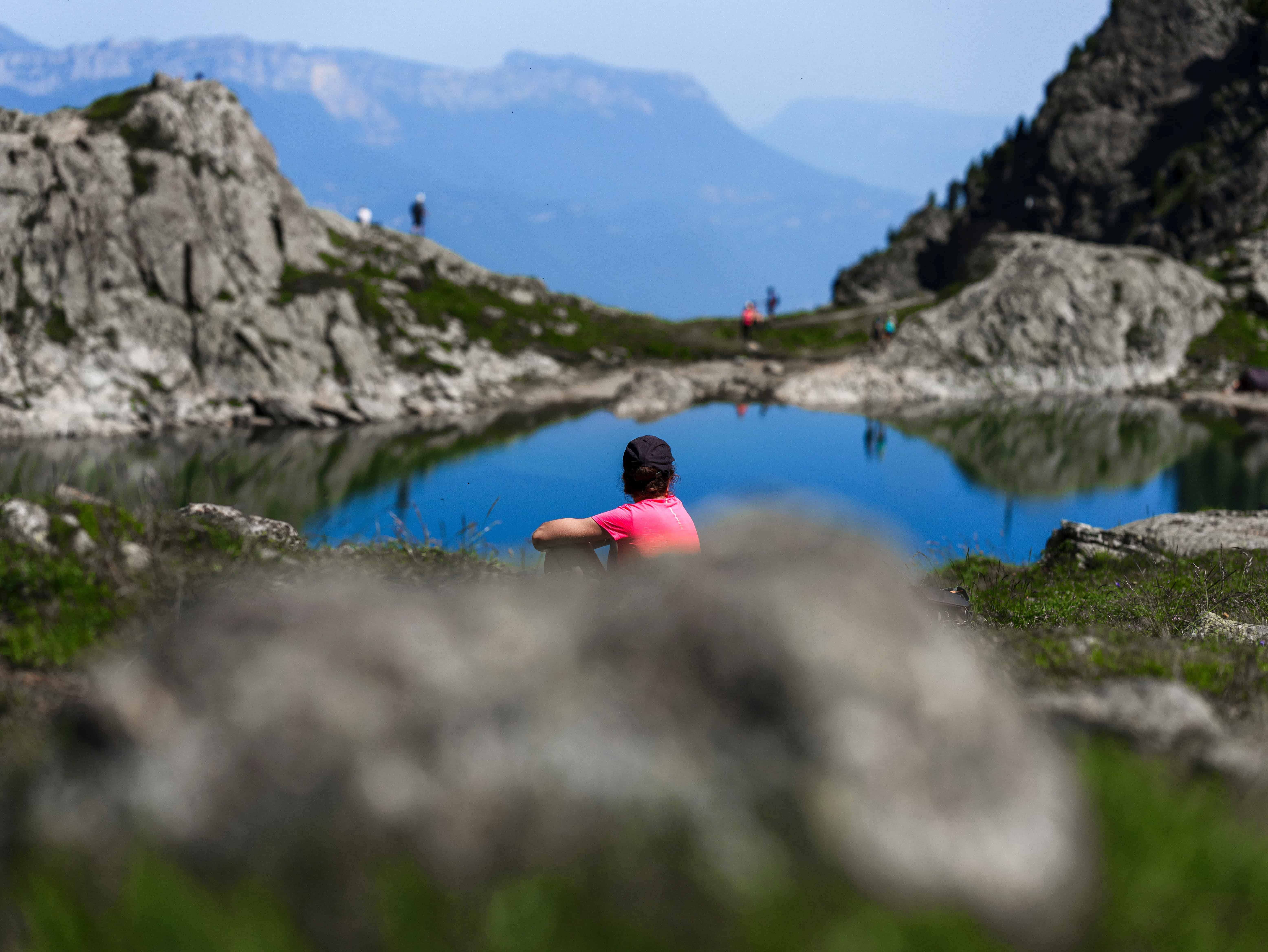 A person in a red shirt sits next to Arrowhead Lake which is surrounded by mountains