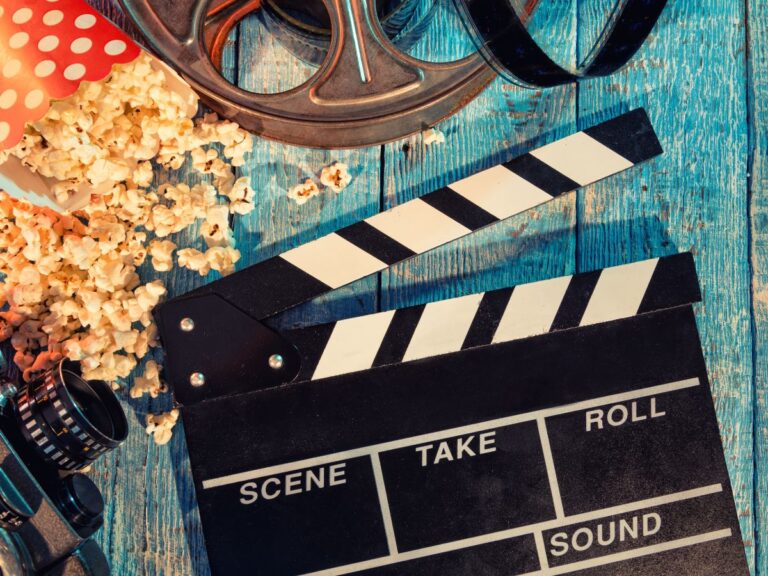 A blue wooden background has a film reel and directors board on it with popcorn in the upper left corner.