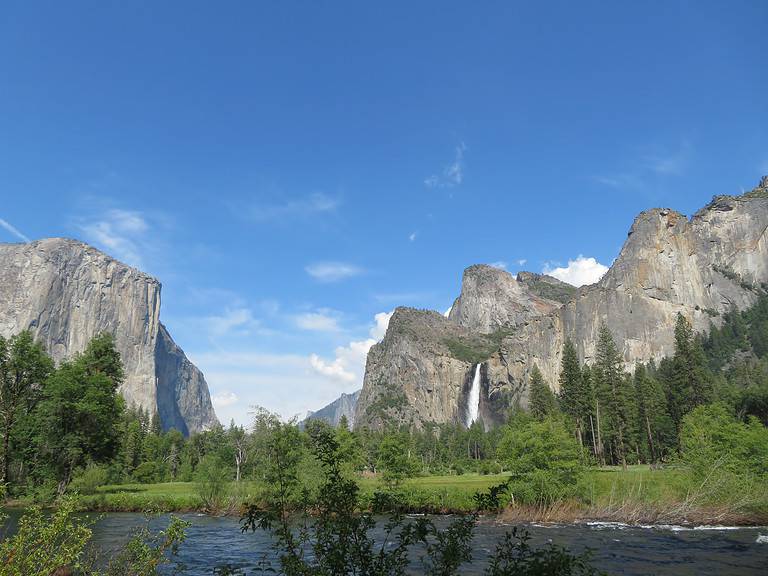 A river runs through a grassy meadow with large mountains on either side.  On the right side there is a large waterfall coming off of the moutnain in Yosemite National PArk.