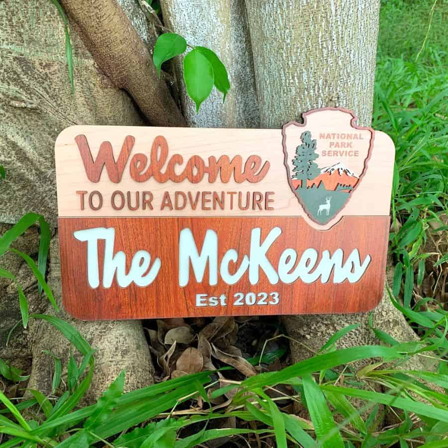 Leaning up against a tree is a wooden sign that resembles the signs found in national parks.  This national park gift can be personalized to include the name of the family.  Writing says. "Welcome to our Adventure"