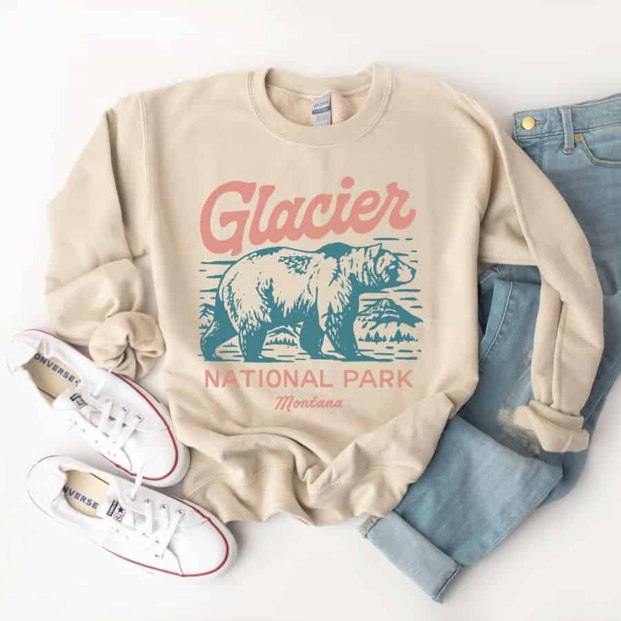 A white long-sleeved t-shirt has the word "Glacier" in pink letters above a graphic of a blue grizzly bear.