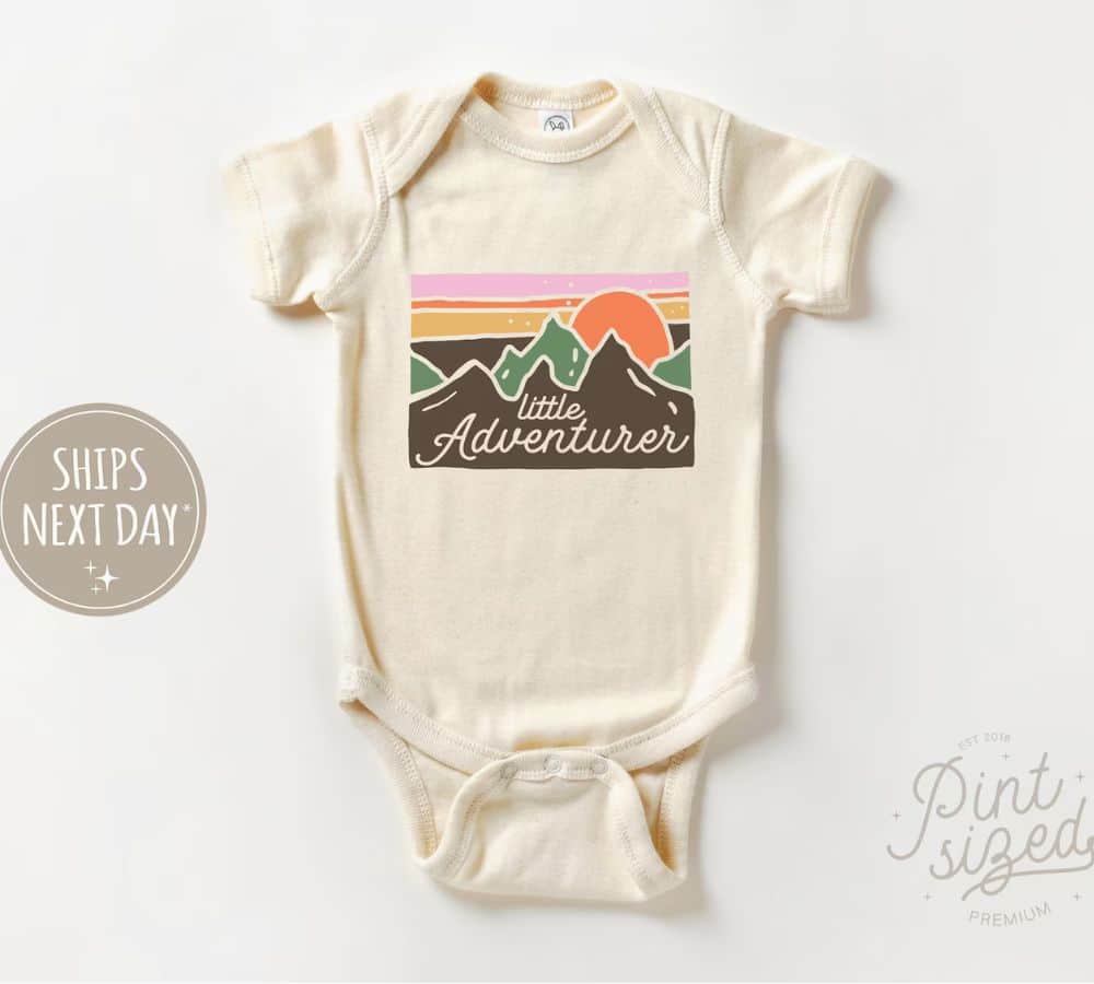 A cream colored onsie has brown mountains with the sun setting over top.  In orange lettering it says "little adventurer"/