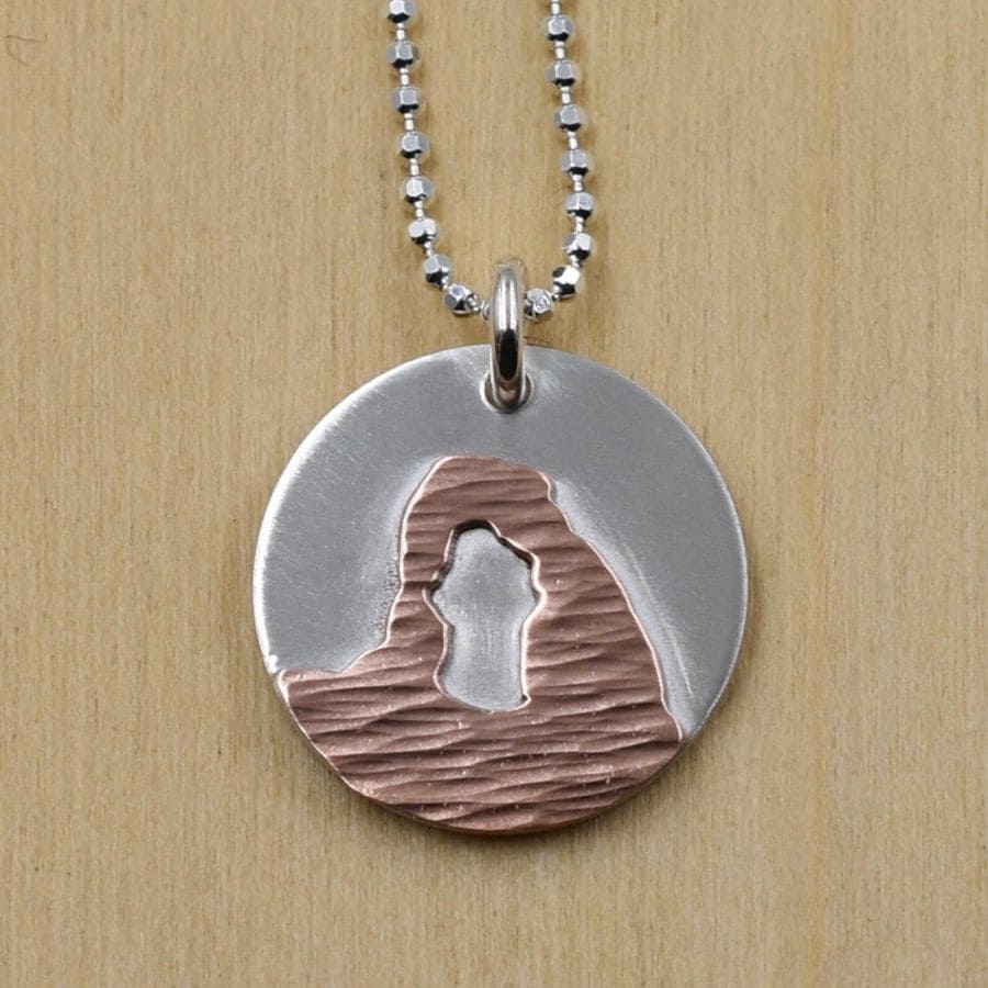 Set on a gold colored background is a round silver necklace with a picture of delicate arch on it.