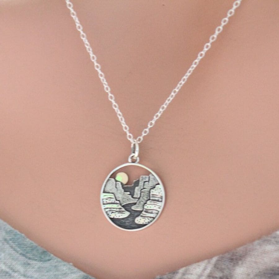 A round silver necklace hangs on a women's neck and contains a die cut of a canyon biew with the sun over top.