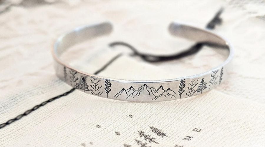 A silver cuff bracelet has etchings of mountains and trees around it.