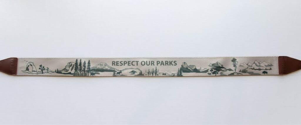 A camera strap is streched out horizontally and has green graphics of a nature scene.  In green letters it says "Protect Our Parks"