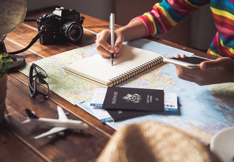 A white woman is sitting at a wooden desk and writing in a notebook.  She has a map, her phone, a black camera, two passports, glasses, and model airplane on the desk as she plans her next trip.  Instead of doing this work yourself, you can purchase my itinerary planning services to do it for you.