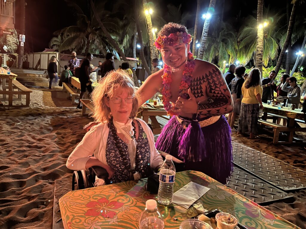 A blonde woman sits in a wheelchair at a luau in Hawaii wearing a blue sundress with white flowers on it.