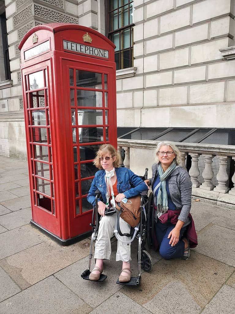 A red telephone booth sits on the sidewalk in London.  Me (a blonde white woman in a wheelchair) and a friend are posing next to the booth.  I'm wearing khaki pants, an orange t-shirt and a blue jean jacket that was part of my London trip packing list.