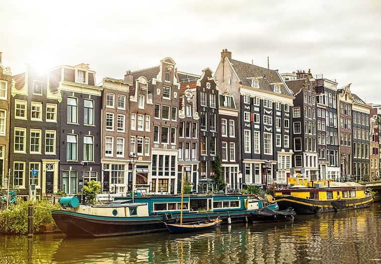 Two houseboats are docked along an Amsterdam canal in front of gingerbread style buildings.