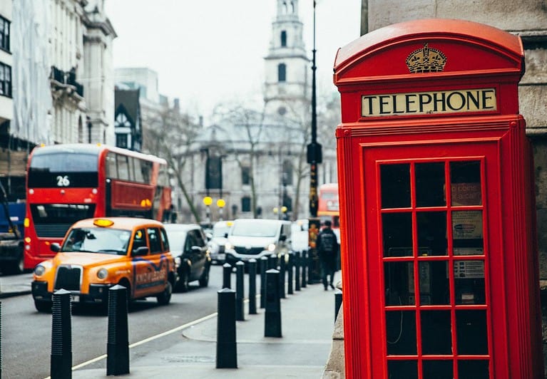 On the corner of a London sidewalk is a red telephone booth.  On the left side of the picture is a busy London street in winter with the trees devoid of leaves.