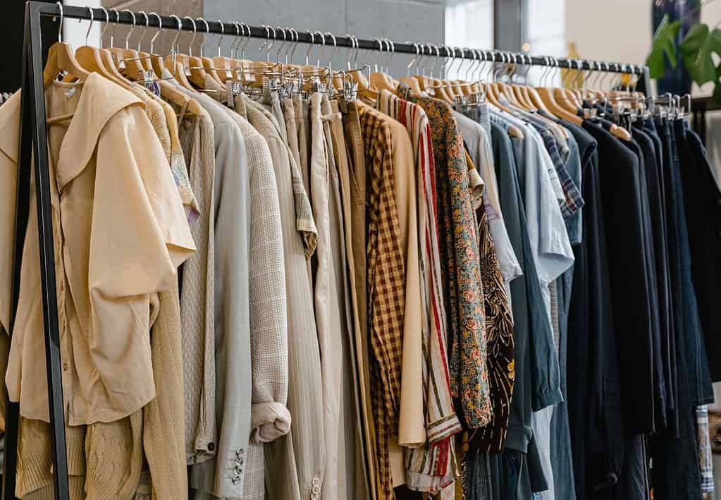 A free standing clothes rack holds a large number of garments including shirts, jackets, blazers, jeans, and other pants in a variety of shades of brown, beige, and blue.  A capsule wardrobe like this is a wonderful addition to your London trip packing list.