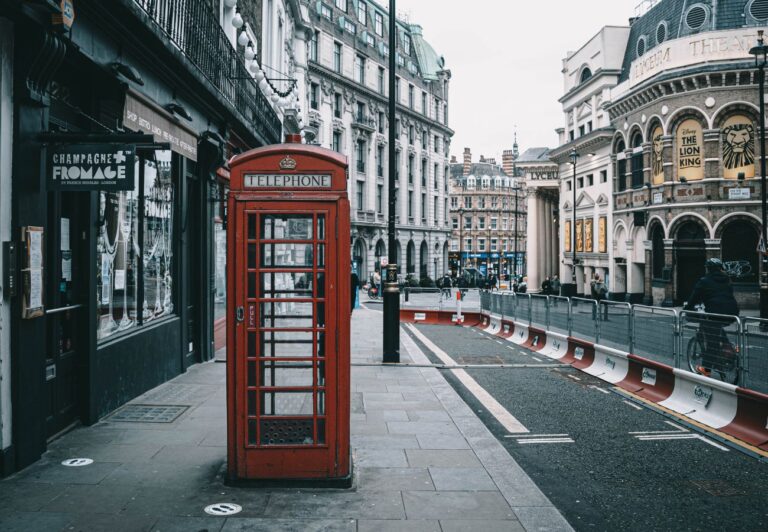 A red telephone booth is in the middle of a sidewalk in London.  There is a theater across the street with Lion King musical posters in the windows.  It's a rainy overcast day.  October is one of the worst times to visit London due to it being the rainiest month.