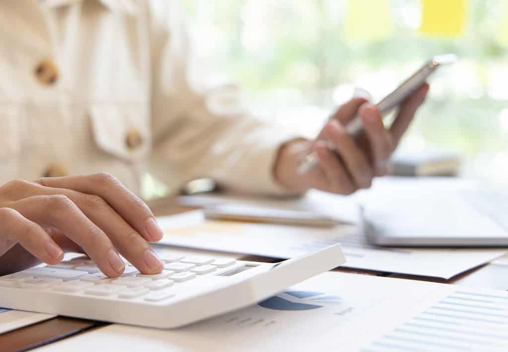 A white person is sitting at a desk using a white calculator and holding a silver cell phone trying to calculate a trip budget.