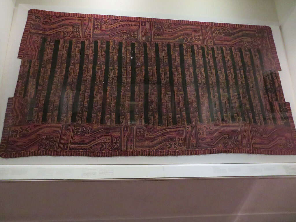 A red and black woven tapestry that was created by the Incas hangs behind glass in a museum