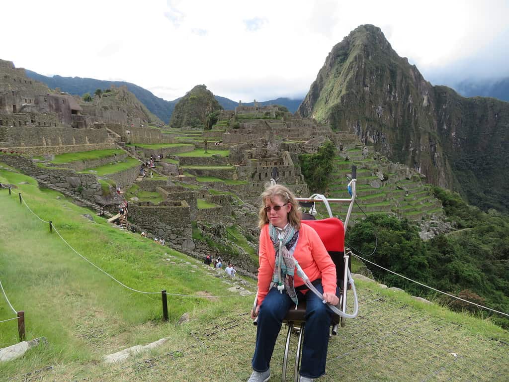 A blonde white woman sits in an adapted one wheeled wheelchair at an overlook with Machu Picchu in the background.