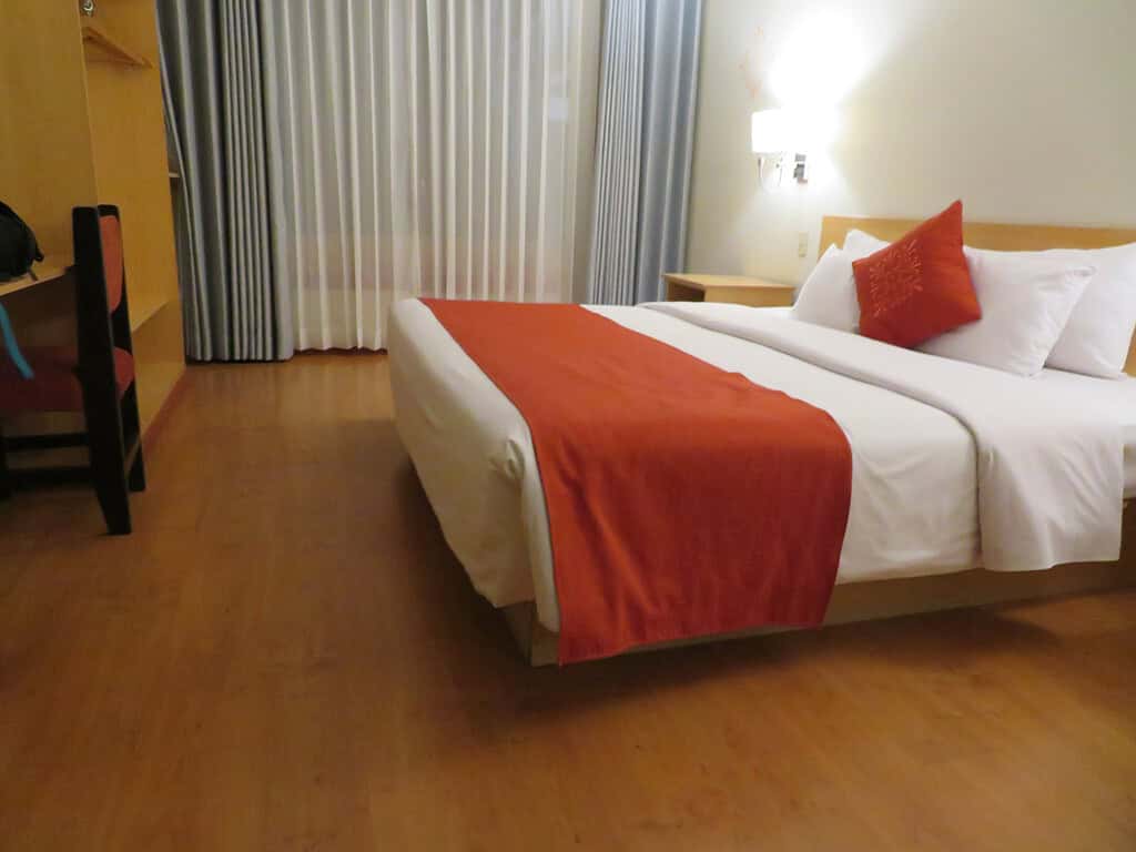 A spacious hotel room is large enough for a wheelchair to maneuver in it and has a large bed with orange accents.