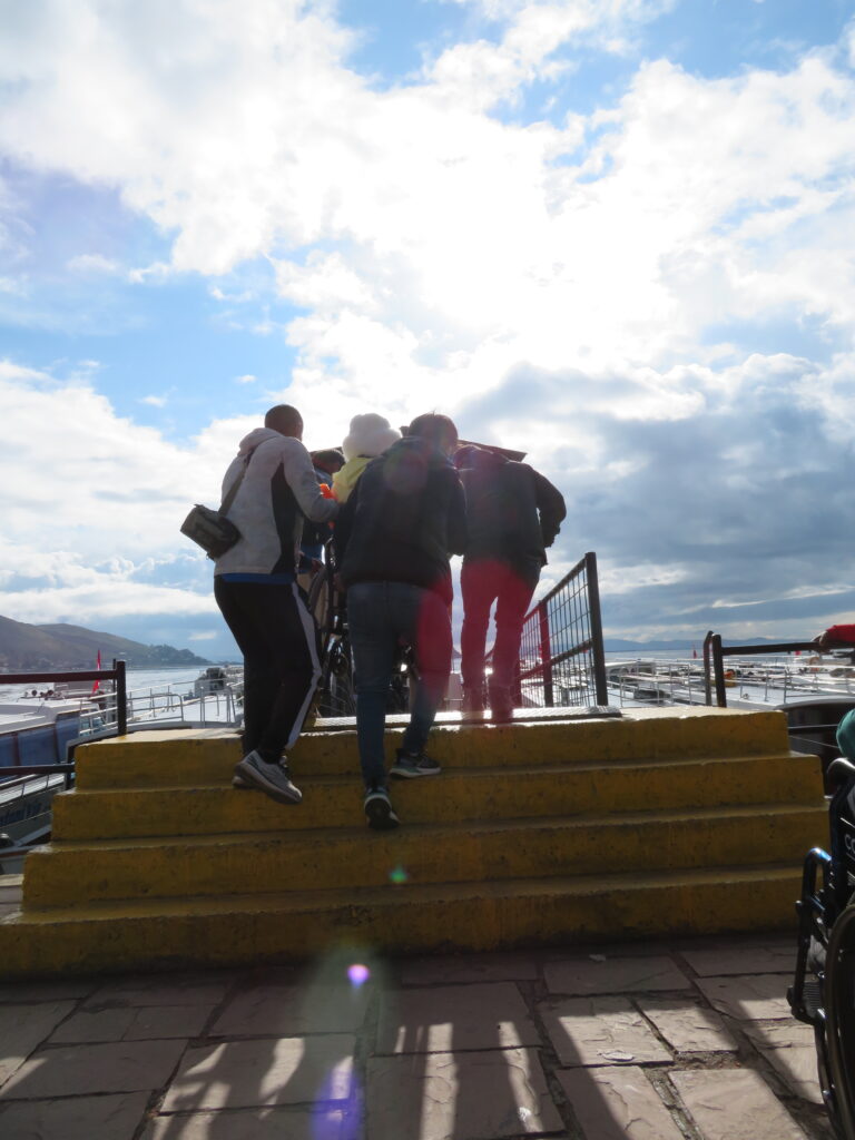 Three men carry a woman in a wheelchair up 4 stairs so that she can get to the boarding area of a boat on lake Titicaca in Peru.