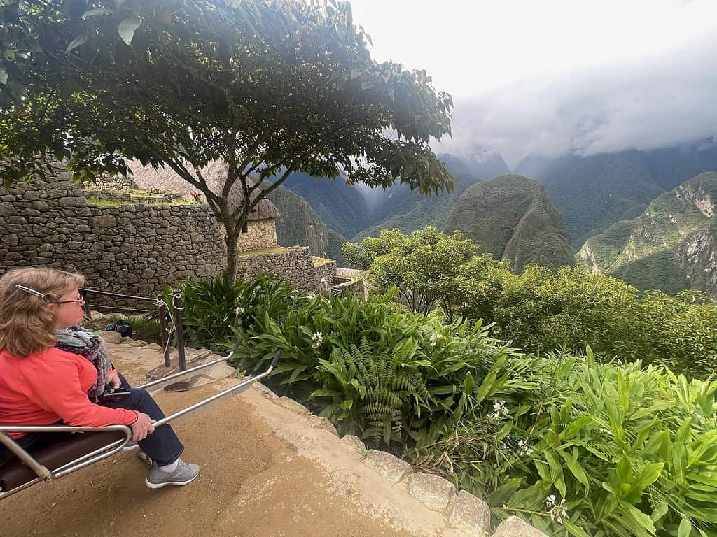 A blonde haired white woman (me) is sitting in an adapted wheelchair on the side of a cliff overlooking the mountains that surround Machu Picchu