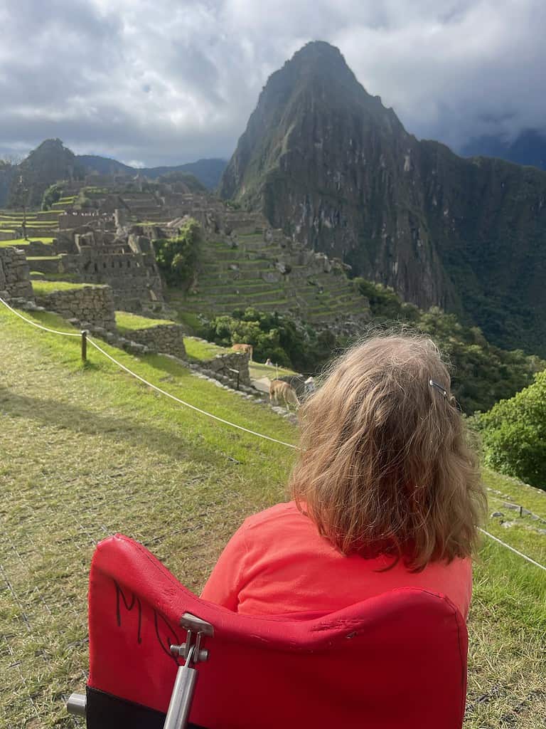 A blonde haired woman is sitting in an adapted wheelchair near the side of a cliff on a mountainside overlooking the ancient Incan site of Machu Picchu in Peru.