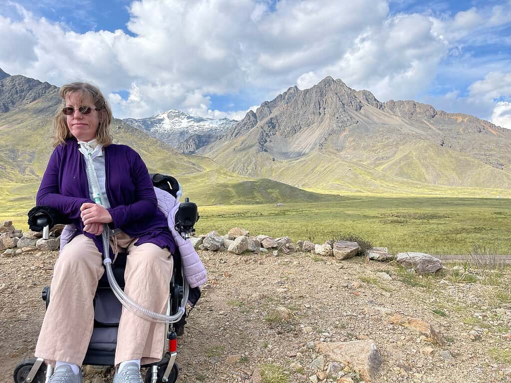 A blonde haired woman in a purple shirt and khaki pants sits in a wheelchair on a gravel area at the base of three mountains in Peru.