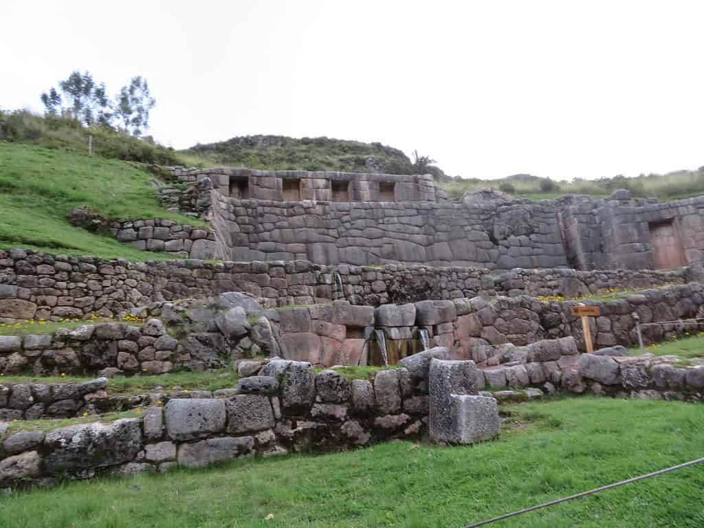 Stone terraced walls are built into the side of a hill and have waterfalls coming out of the lower level.  This is the Inca site of Tambomachay.  Sites like these are worth visiting during your 8 days in Peru.