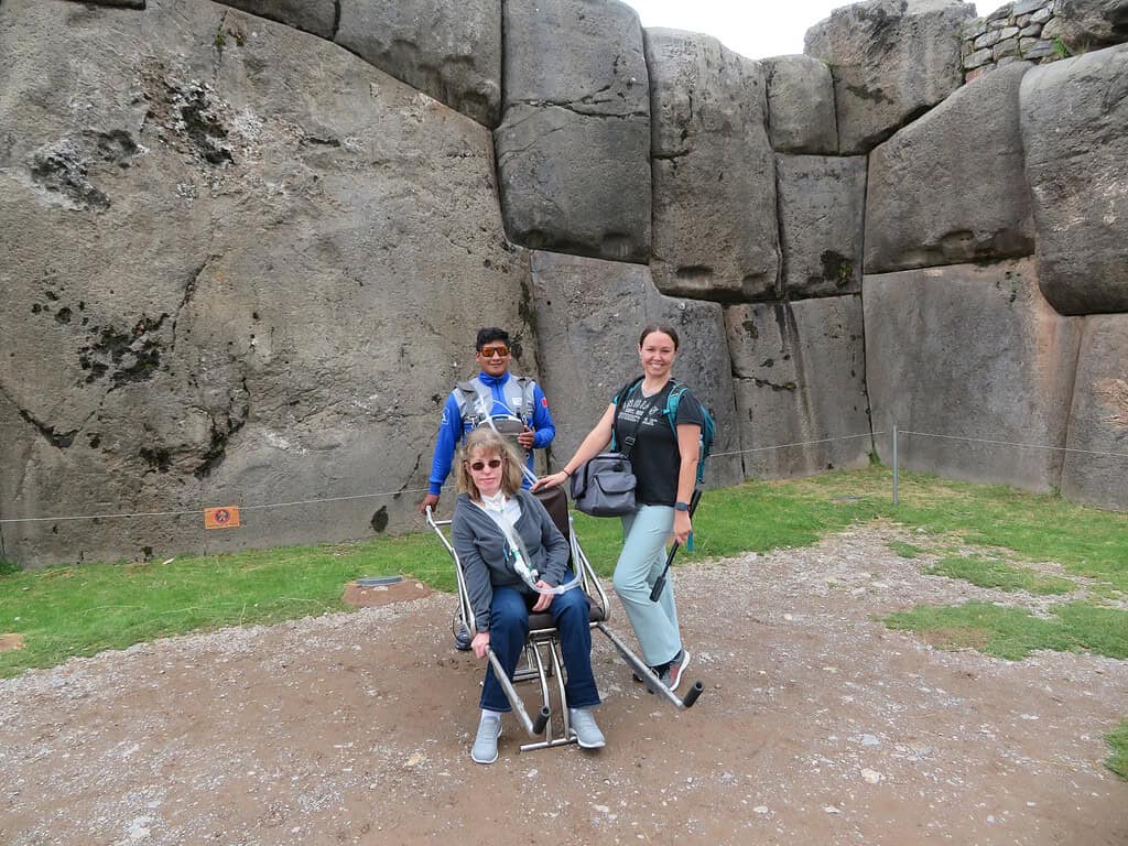 I am a blonde haired woman in an adapted wheelchair at the base of Saqsaywaman with another white woman with brown hair and a Peruvian man with black hair standing with me.