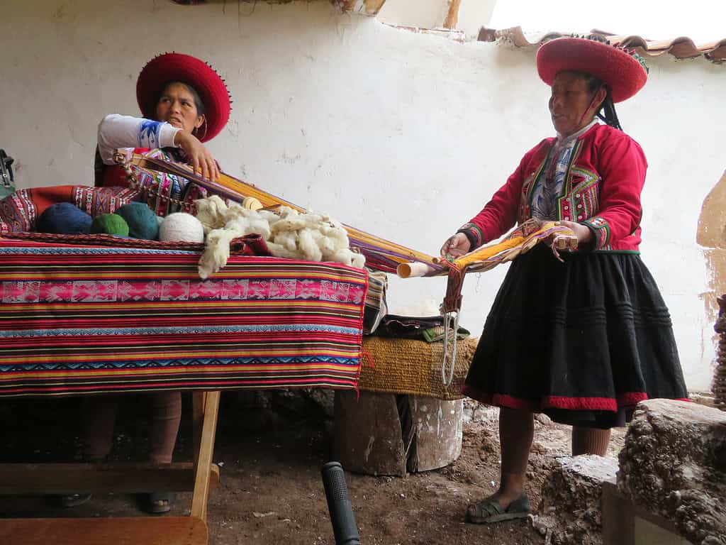 Two Peruvian women with black braided hair wear red jackets and black skirts.  They are demonstrating the weaving process where they make handmade tavle runners and other textiles.