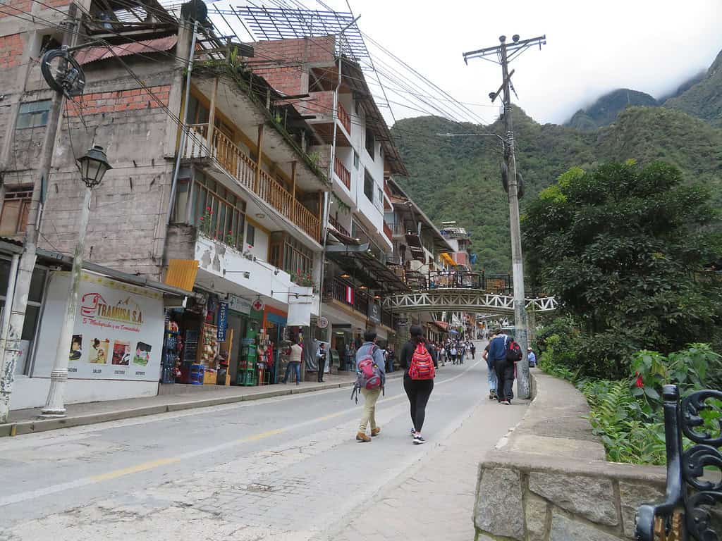 The main street of Aguas Calientes near Machu Picchu is at the base of a mountain range and has white buildings on the left hand side of the street.