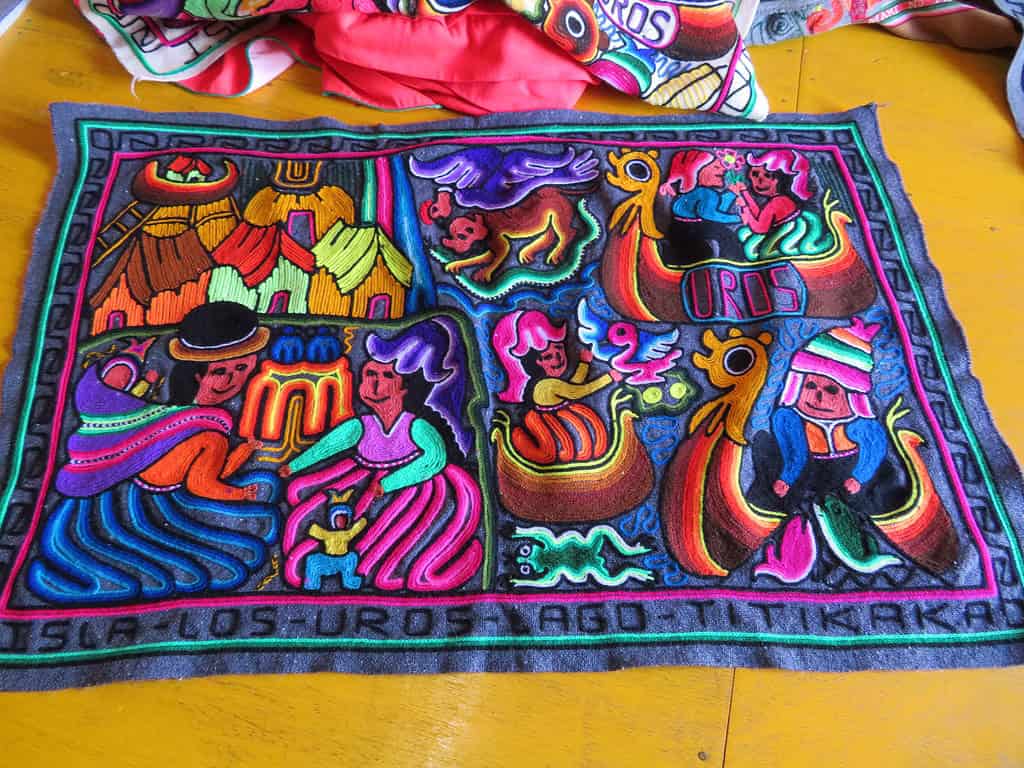 A handmade woven textile has bright colors of pink, red, purple, and teal and depicts life on the islands of Uros.