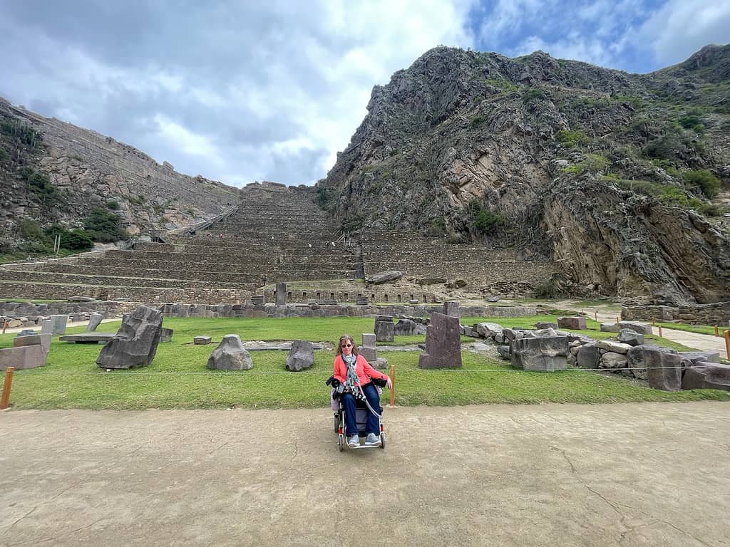 A white woman with blonde hair is wearing a pink sweater and blue jeans and is sitting in a wheelchair at the base of the old Incan site of Ollantaytambo.