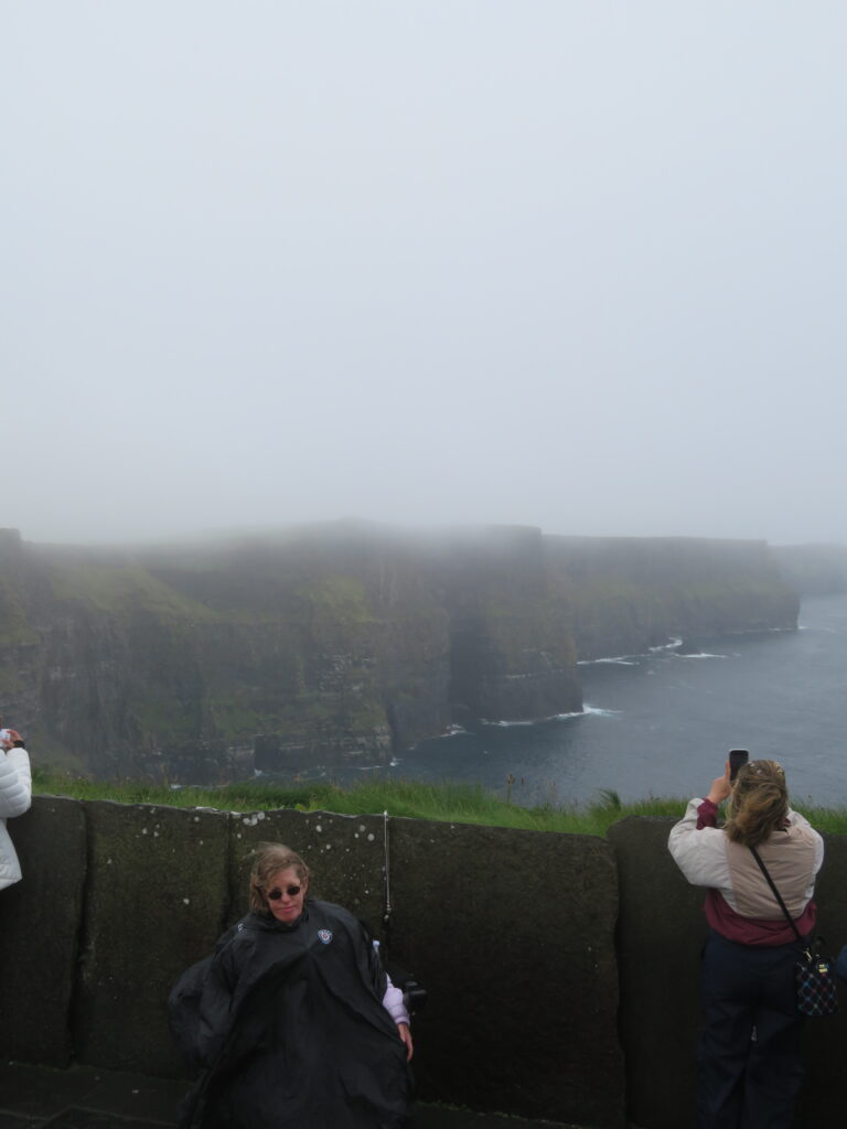 A blonde woman in a wheelchair sits in front of a sonte barrier along the Cliffs of Moher on a foggy day.  The view of the cliffs are obstruvted to wheelchair users due to the stone walls that act as barriers and may make a visit to the cliffs of Moher not worth it.