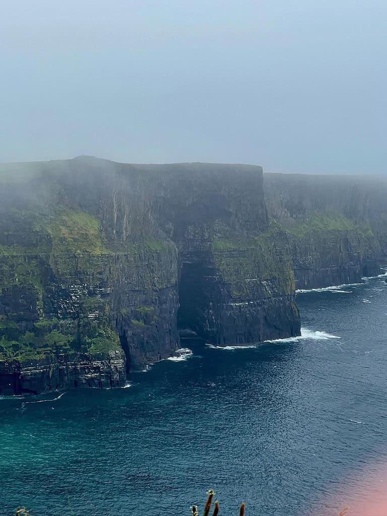 A large stone cliff has a natrually formed cave at its base and is part of the Cliffs of Moher in Ireland.  Fog surrounds the cliffs for an eerie view.