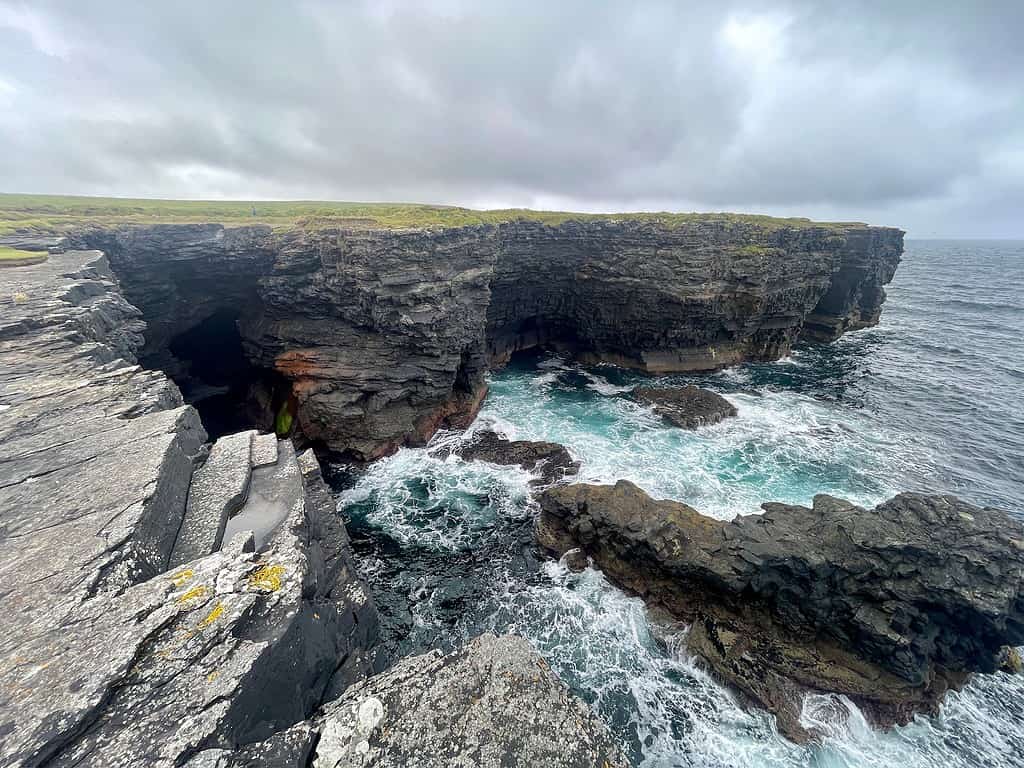 Water crashes against rocky cliffs and form a natural cave.  These tall stone outcrops have green grass on top of them and are known as Kilkee Cliffs in Ireland