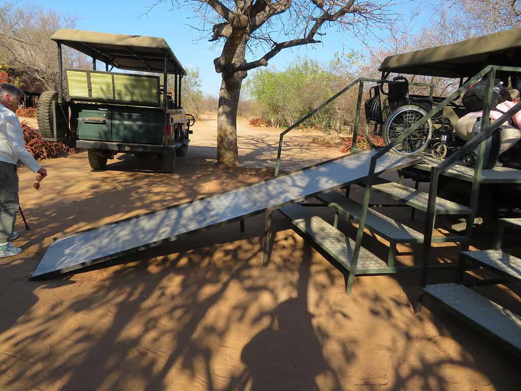 A high clearance open air jeep is parked in front of a set of 4 stairs which allow people to enter it more easily.  A steep ramp goes up the left side of the stairs to allow access for wheelchairs.