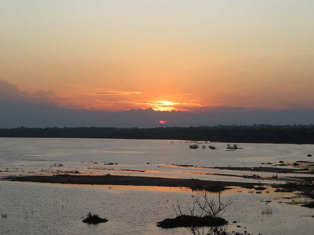 The sun sets over a river in Uganda creating yellow, orange, and red colors in the sky which reflect in the river below.